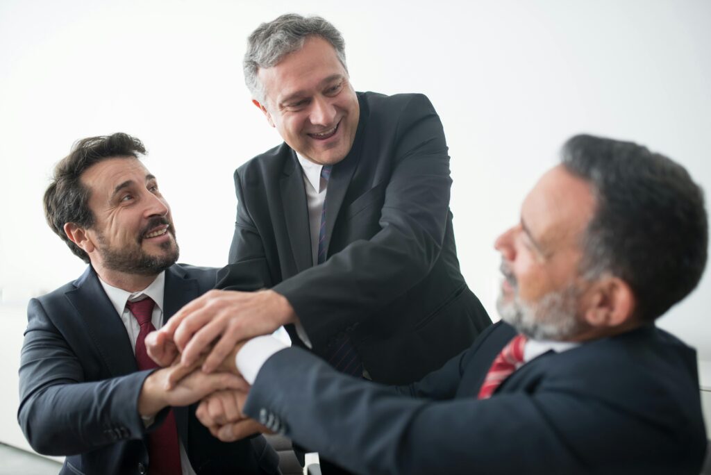 Three businessmen in suits celebrating teamwork success indoors with handshake.